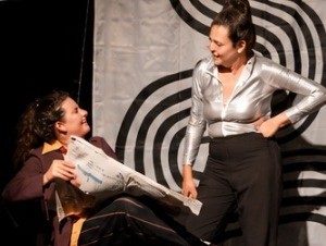 Zwei Schauspielerinnen auf der Bühne, eine liest Zeitung
